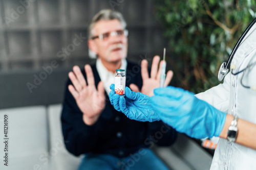 Close up of a nurse s hand in blue protective gloves holding an ampoule with the vaccine and a prick in his hands  on the background a blurred man who is saying the vaccine. Vaccine against covid-19. 