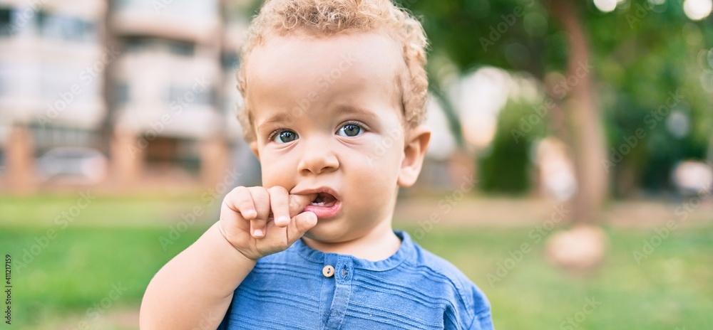 Sad little boy putting fingers on mouth touching gums because toothache at the park on a sunny day. Beautiful blonde hair male toddler in pain for new baby teeth outdoors Sad little boy putting fingers on mouth touching gums because toothache at the park on a sunny day. Beautiful blonde hair male toddler in pain for new baby teeth outdoors