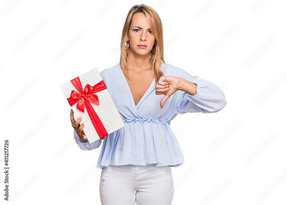 Beautiful blonde woman holding gift with angry face, negative sign showing dislike with thumbs down, rejection concept Beautiful blonde woman holding gift with angry face, negative sign showing dislike with thumbs down, rejection concept