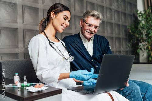A smiling nurse visits an older man's home, she points a finger at the computer about the test results. Doctor's visit home, remote treatment at home