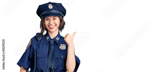 Young beautiful girl wearing police uniform smiling with happy face looking and pointing to the side with thumb up.