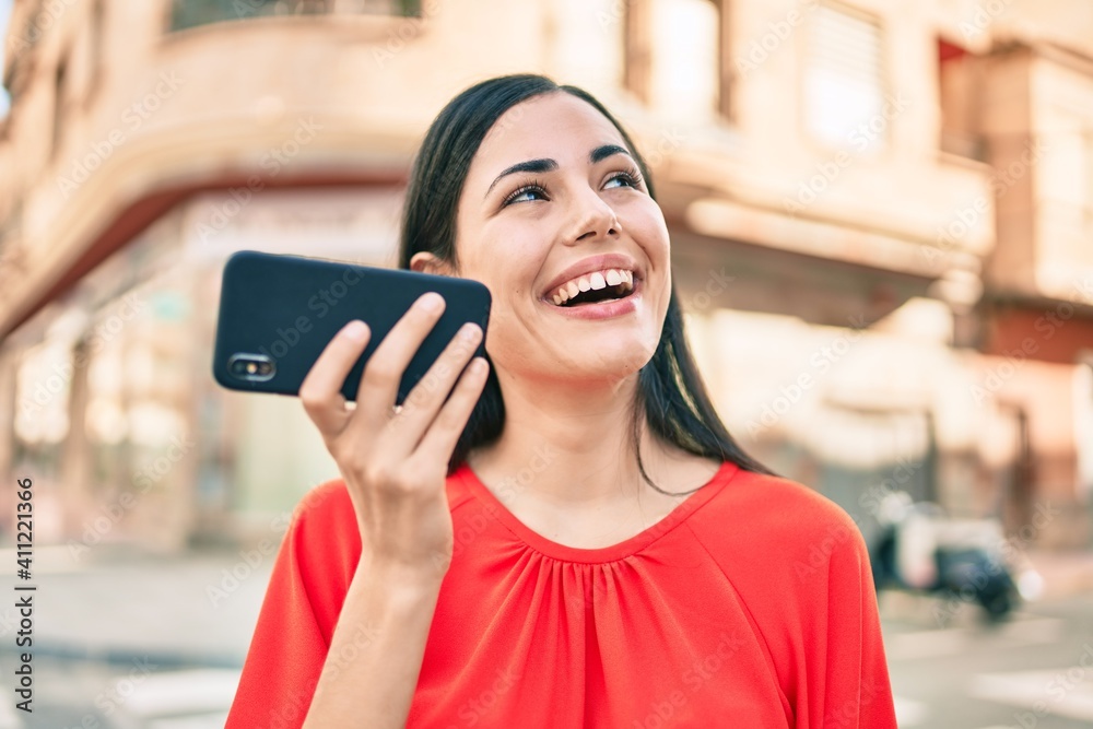 Young latin girl smiling happy sending voice message using smartphone at the city.