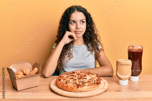 Teenager hispanic girl eating pizza and fried chicken thinking concentrated about doubt with finger on chin and looking up wondering