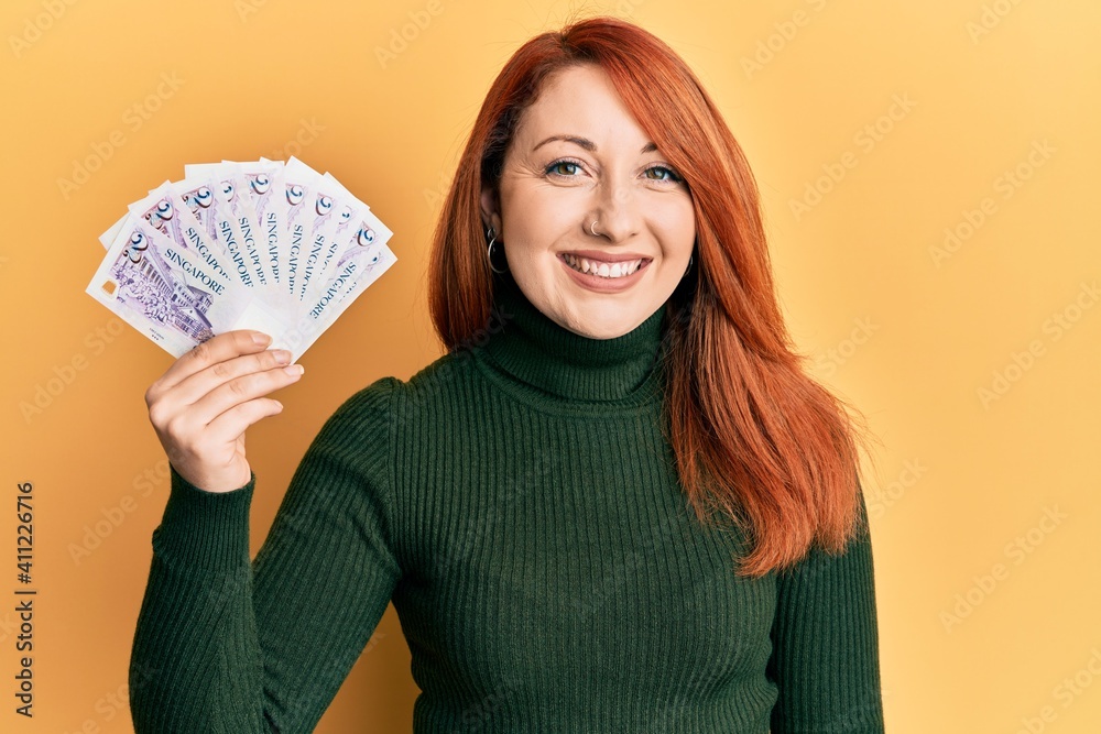 Beautiful redhead woman holding 2 singapore dollars banknotes looking positive and happy standing and smiling with a confident smile showing teeth Beautiful redhead woman holding 2 singapore dollars banknotes looking positive and happy standing and smiling with a confident smile showing teeth