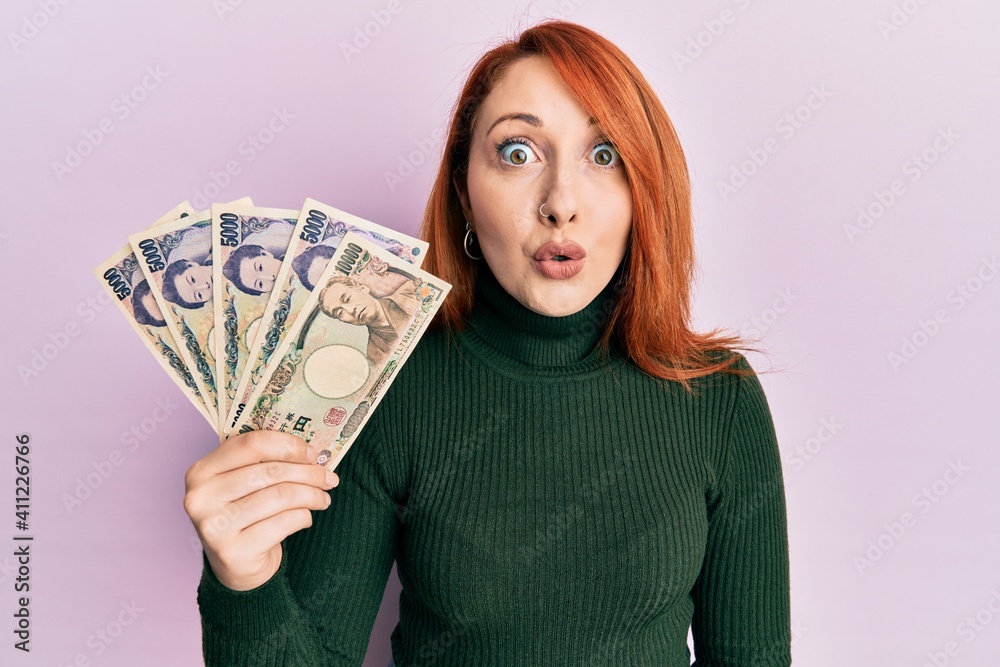 Beautiful redhead woman holding japanese yen banknotes scared and amazed with open mouth for surprise, disbelief face Beautiful redhead woman holding japanese yen banknotes scared and amazed with open mouth for surprise, disbelief face