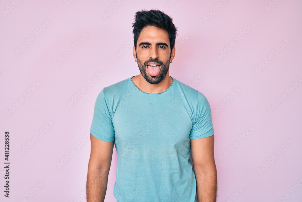 Handsome hispanic man with beard wearing casual clothes sticking tongue out happy with funny expression. emotion concept. Handsome hispanic man with beard wearing casual clothes sticking tongue out happy with funny expression. emotion concept.