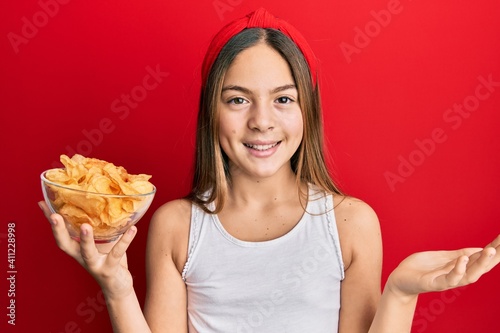 Beautiful brunette little girl holding potato chips celebrating achievement with happy smile and winner expression with raised hand