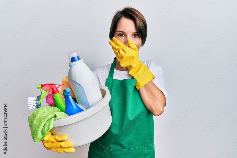 Young brunette woman with short hair wearing apron holding cleaning products shocked covering mouth with hands for mistake. secret concept. Young brunette woman with short hair wearing apron holding cleaning products shocked covering mouth with hands for mistake. secret concept.