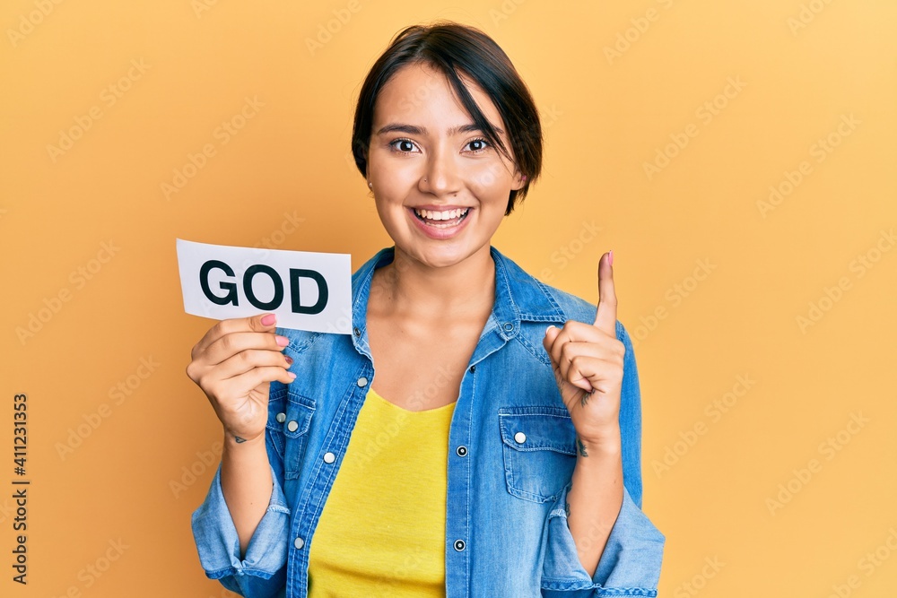 Beautiful young woman with short hair holding paper with god word smiling with an idea or question pointing finger with happy face, number one Beautiful young woman with short hair holding paper with god word smiling with an idea or question pointing finger with happy face, number one