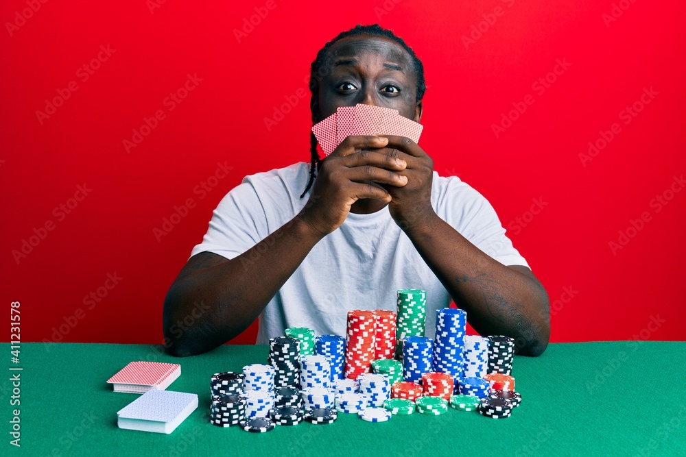 Handsome young black man playing gambling poker covering face with cards puffing cheeks with funny face. mouth inflated with air, catching air. Handsome young black man playing gambling poker covering face with cards puffing cheeks with funny face. mouth inflated with air, catching air.