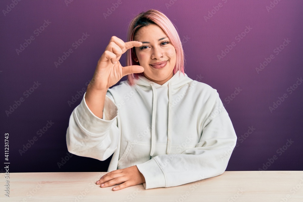 Hispanic woman with pink hair wearing casual sweatshirt sitting on the table smiling and confident gesturing with hand doing small size sign with fingers looking and the camera. measure concept. Hispanic woman with pink hair wearing casual sweatshirt sitting on the table smiling and confident gesturing with hand doing small size sign with fingers looking and the camera. measure concept.