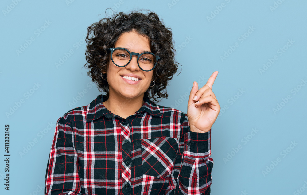Young hispanic woman with curly hair wearing casual clothes and glasses smiling happy pointing with hand and finger to the side Young hispanic woman with curly hair wearing casual clothes and glasses smiling happy pointing with hand and finger to the side