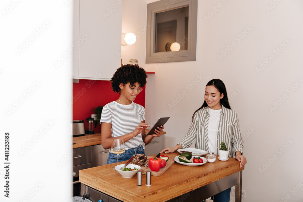 Joyful female friends are standing next to kitchen table with vegetables and smiling