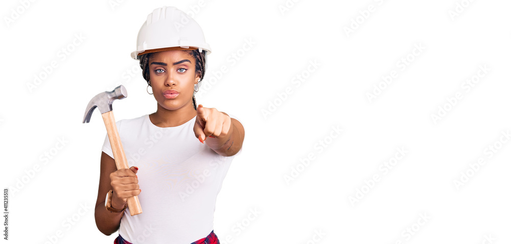 Young african american woman with braids wearing hardhat holding hammer pointing with finger to the camera and to you, confident gesture looking serious Young african american woman with braids wearing hardhat holding hammer pointing with finger to the camera and to you, confident gesture looking serious