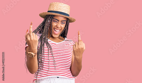 Young african american woman with braids wearing summer hat showing middle finger doing fuck you bad expression, provocation and rude attitude. screaming excited