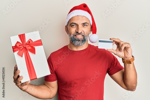 Middle age handsome man wearing chrismast hat holding gift and credit card smiling with a happy and cool smile on face. showing teeth. photo