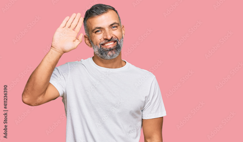 Fototapeta premium Middle age handsome man wearing casual white tshirt waiving saying hello happy and smiling, friendly welcome gesture