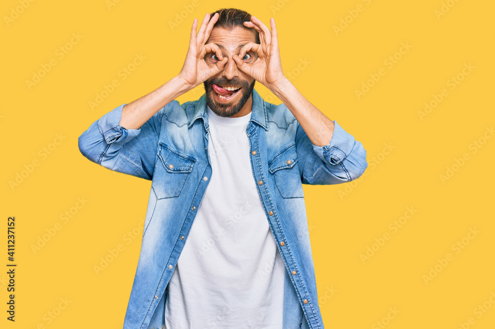 Attractive man with long hair and beard wearing casual denim jacket doing ok gesture like binoculars sticking tongue out, eyes looking through fingers. crazy expression.