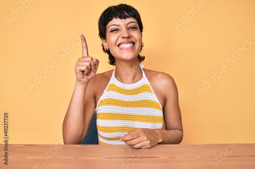 Beautiful brunettte woman sitting on the table over yellow background pointing finger up with successful idea. exited and happy. number one.
