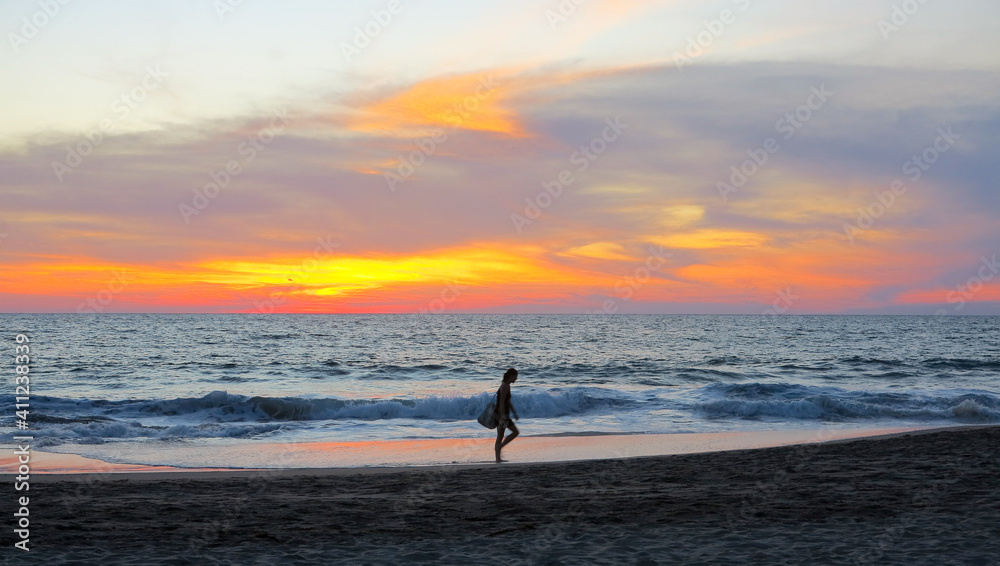 Puerto Escondito, Mexico - December 28 2018 - A woman walking down the beach in front of a beautiful red and orange sunset over the Pacific Ocean in Puerto Escondito, Mexico.  Image has copy space.