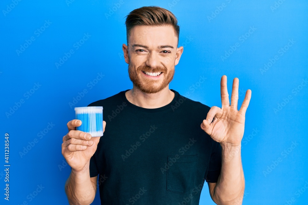 Young redhead man holding earwax cotton remover doing ok sign with fingers, smiling friendly gesturing excellent symbol Young redhead man holding earwax cotton remover doing ok sign with fingers, smiling friendly gesturing excellent symbol