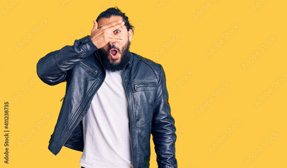 Young arab man wearing casual leather jacket peeking in shock covering face and eyes with hand, looking through fingers with embarrassed expression. Young arab man wearing casual leather jacket peeking in shock covering face and eyes with hand, looking through fingers with embarrassed expression.