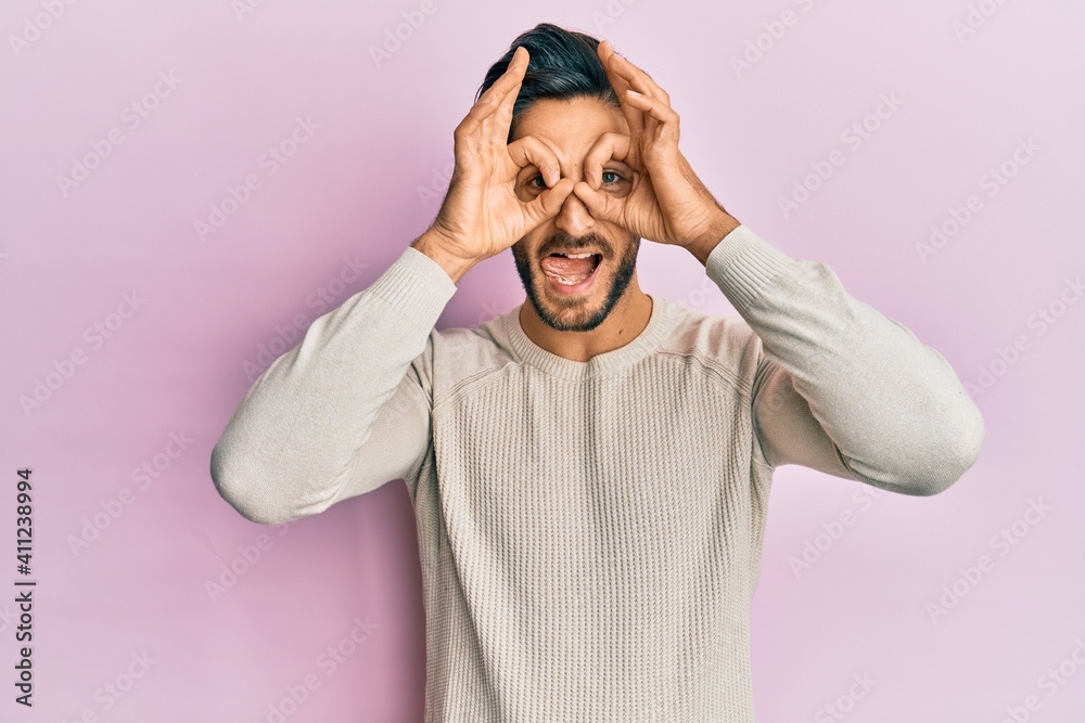Young hispanic man wearing casual winter sweater doing ok gesture like binoculars sticking tongue out, eyes looking through fingers. crazy expression. Young hispanic man wearing casual winter sweater doing ok gesture like binoculars sticking tongue out, eyes looking through fingers. crazy expression.