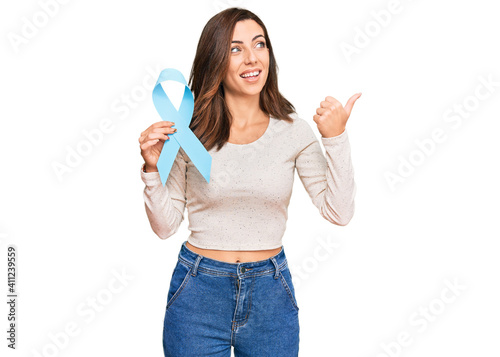 Young brunette woman holding blue ribbon pointing thumb up to the side smiling happy with open mouth