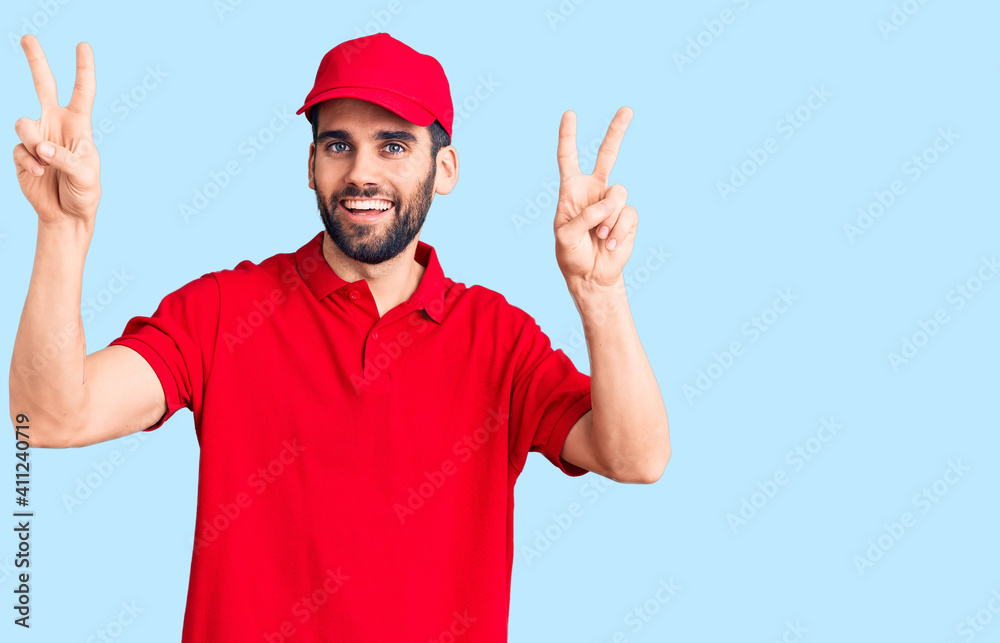 Young handsome man with beard wearing delivery uniform smiling with tongue out showing fingers of both hands doing victory sign. number two.