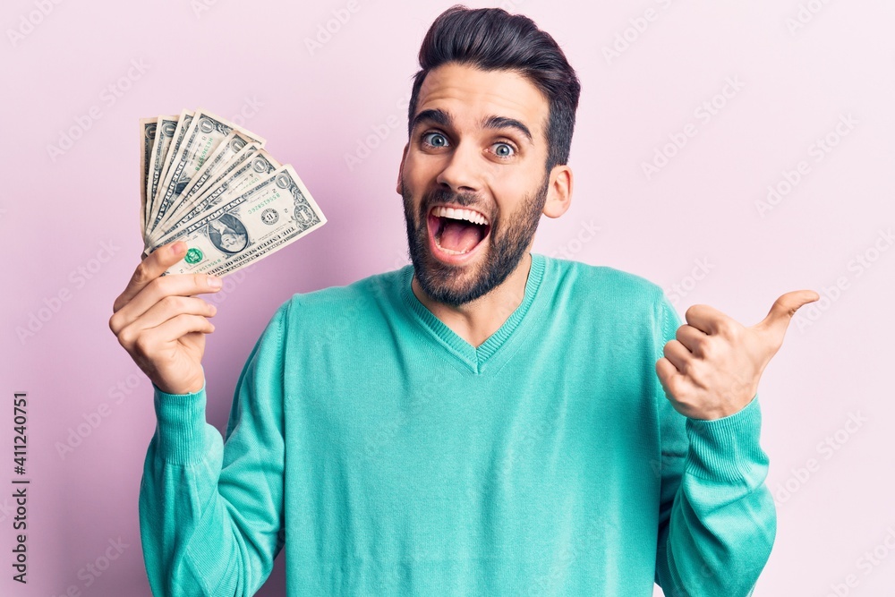 Young handsome man with beard holding bunch of dollar pointing thumb up to the side smiling happy with open mouth Young handsome man with beard holding bunch of dollar pointing thumb up to the side smiling happy with open mouth
