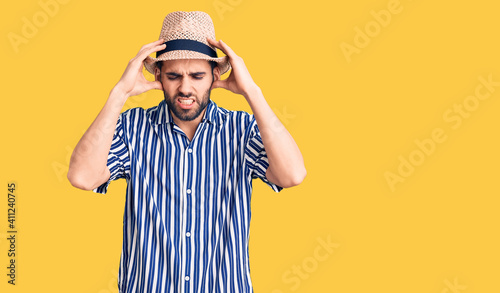Young handsome man with beard wearing summer hat and striped shirt with hand on head for pain in head because stress. suffering migraine.