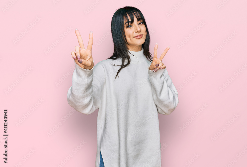 Young brunette woman with bangs wearing casual turtleneck sweater smiling looking to the camera showing fingers doing victory sign. number two.