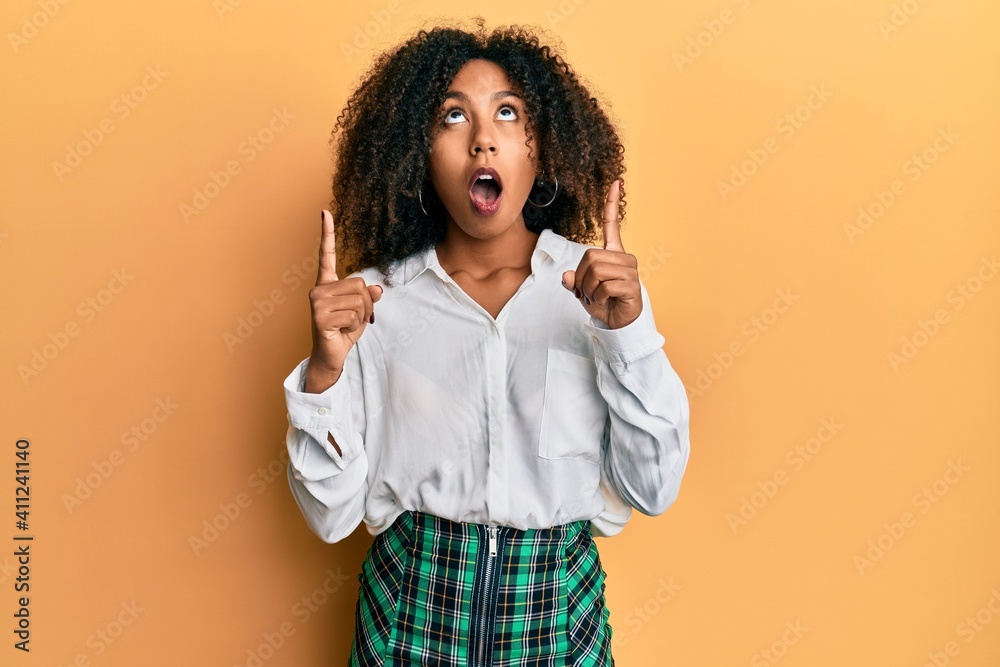 Beautiful african american woman with afro hair wearing scholar skirt amazed and surprised looking up and pointing with fingers and raised arms.