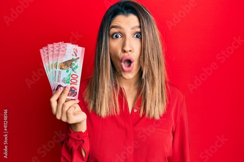 Beautiful brunette woman holding 100 new zealand dollars banknote scared and amazed with open mouth for surprise, disbelief face