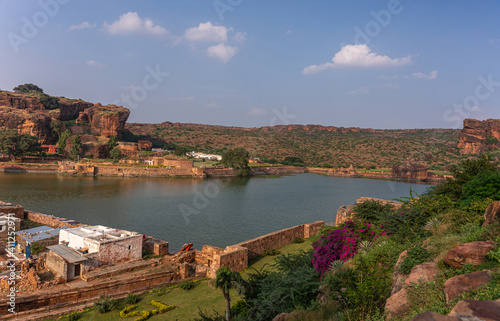 Badami, Karnataka, India - November 7, 2013: Cave temples above Agasthya Lake as shown, surrounded by hills, rocks, and cliffs under blue cloudscape. Flowers add color, Green foliage.