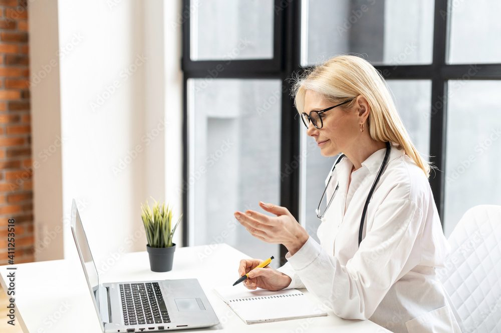 Confident and helpful middle-aged MD, pediatric, physician wearing white medical gown in modern clinic office and talks with patient via video connection on the laptop, consults online. Side view Confident and helpful middle-aged MD, pediatric, physician wearing white medical gown in modern clinic office and talks with patient via video connection on the laptop, consults online. Side view