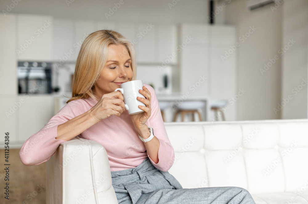 A calm and peaceful middle-aged woman takes a break, rests with a cup of hot tea on the couch at home. Contented charming mature lady enjoying coffee and daydreaming A calm and peaceful middle-aged woman takes a break, rests with a cup of hot tea on the couch at home. Contented charming mature lady enjoying coffee and daydreaming