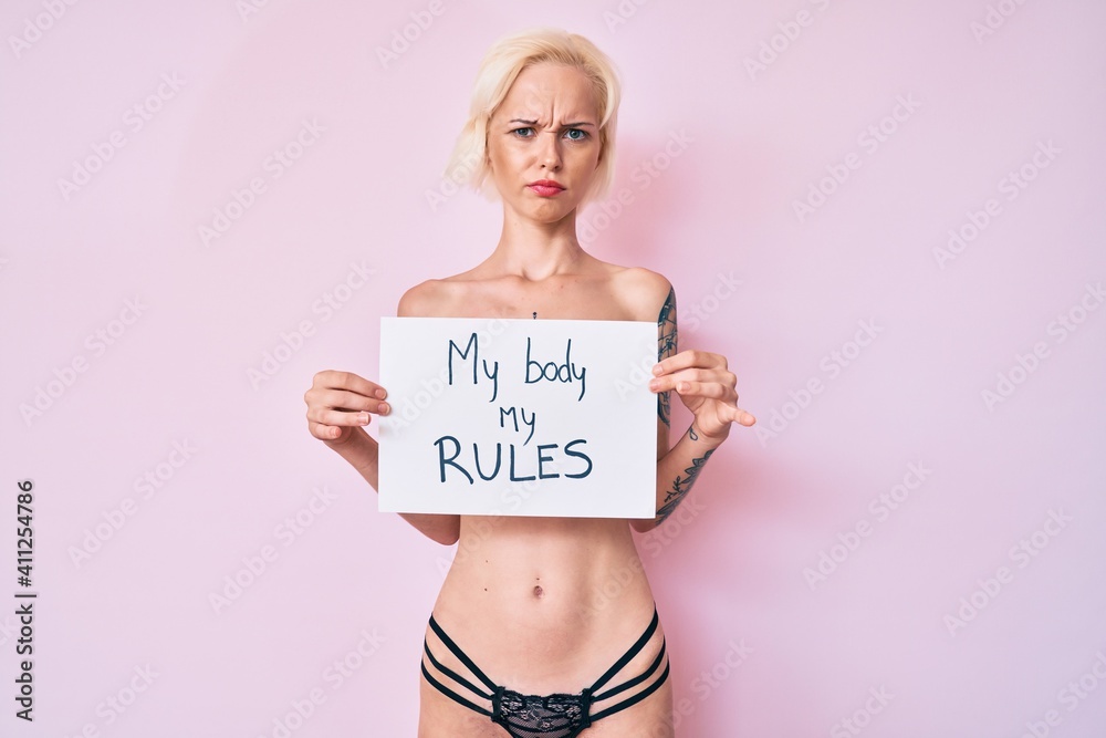 Young blonde woman with tattoo standing shirtless holding banner with my body my rules message skeptic and nervous, frowning upset because of problem. negative person. Young blonde woman with tattoo standing shirtless holding banner with my body my rules message skeptic and nervous, frowning upset because of problem. negative person.