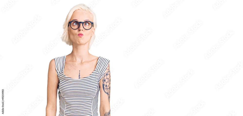 Young blonde woman with tattoo wearing casual clothes and glasses making fish face with lips, crazy and comical gesture. funny expression. Young blonde woman with tattoo wearing casual clothes and glasses making fish face with lips, crazy and comical gesture. funny expression.