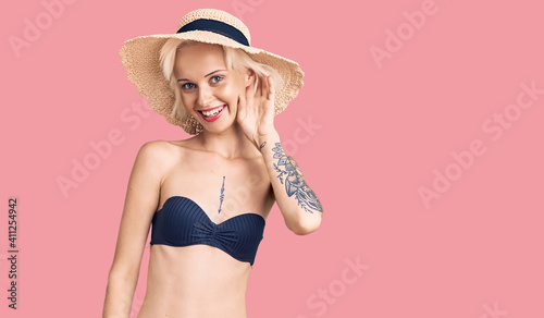 Young blonde woman with tattoo wearing bikini and summer hat smiling with hand over ear listening an hearing to rumor or gossip. deafness concept.