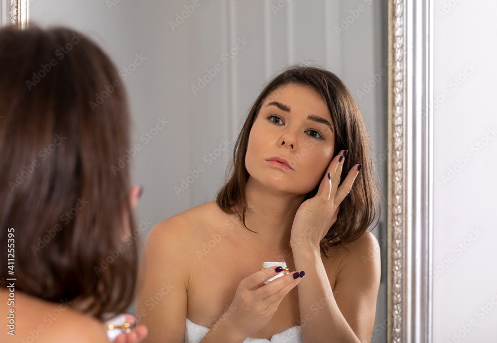 Obraz premium young beautiful woman with dark hair applies cream to her face in front of a mirror, problem aged skin concept, serious face
