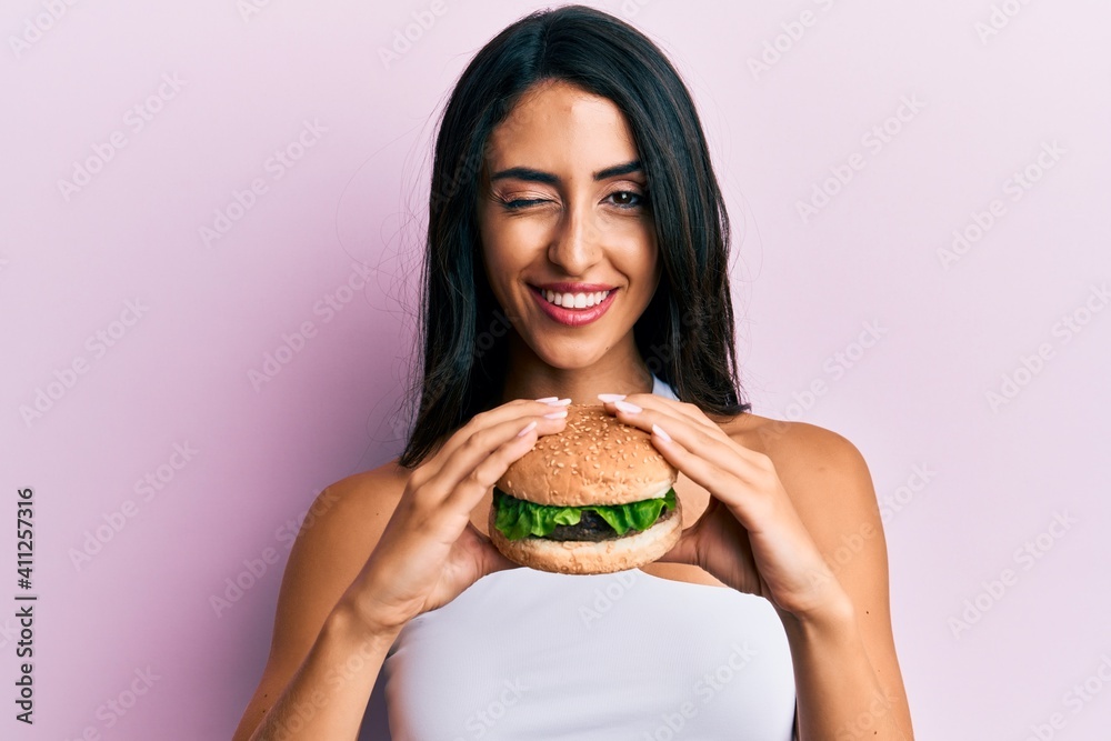 Beautiful hispanic woman eating a tasty classic burger winking looking at the camera with sexy expression, cheerful and happy face.