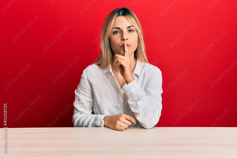 Young caucasian woman wearing casual clothes sitting on the table asking to be quiet with finger on lips. silence and secret concept. Young caucasian woman wearing casual clothes sitting on the table asking to be quiet with finger on lips. silence and secret concept.