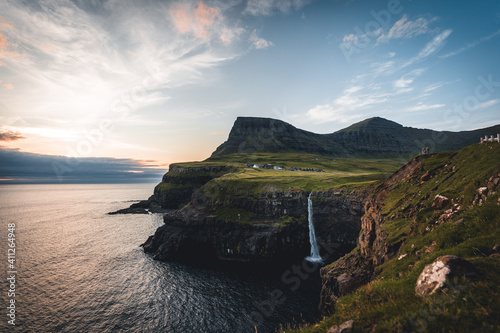 Gasadalur village and Mulafossur its iconic waterfall during summer with bluw sky. Vagar, Faroe Islands, Denmark. Rough see in the north atlantic ocean.