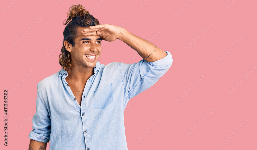 Young hispanic man wearing summer style very happy and smiling looking far away with hand over head. searching concept. Young hispanic man wearing summer style very happy and smiling looking far away with hand over head. searching concept.
