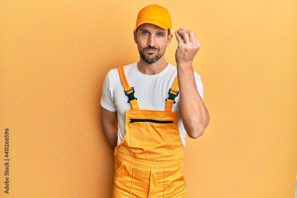 Young handsome man wearing handyman uniform over yellow background doing italian gesture with hand and fingers confident expression
