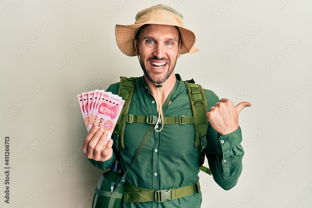 Handsome man with beard wearing explorer hat holding 100 yuan banknotes pointing thumb up to the side smiling happy with open mouth Handsome man with beard wearing explorer hat holding 100 yuan banknotes pointing thumb up to the side smiling happy with open mouth