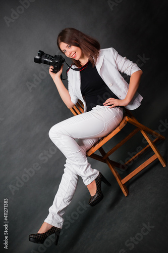 Attractive girl photographer sits on a chair with a camera in her hands in the studio on a gray background.