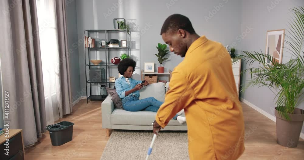 Happy handsome young African American husband mopping the floor in front while pretty wife resting on couch texting on smartphone browsing online social network app, cleaning home concept Happy handsome young African American husband mopping the floor in front while pretty wife resting on couch texting on smartphone browsing online social network app, cleaning home concept
