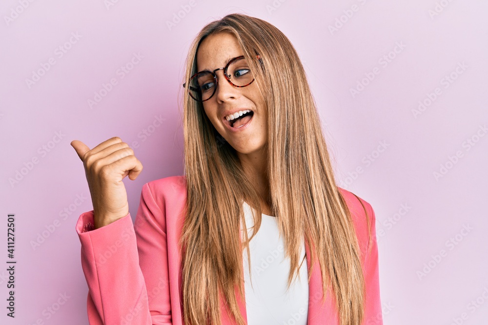 Young blonde woman wearing business style and glasses smiling with happy face looking and pointing to the side with thumb up. Young blonde woman wearing business style and glasses smiling with happy face looking and pointing to the side with thumb up.